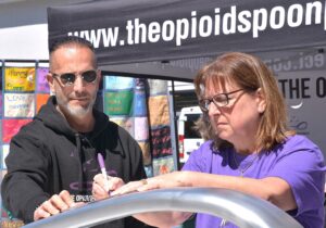 At a 2019 fundraiser for Team Sharing, the Opioid Spoon Project founder and artist/activist Domenic Esposito assists Cheryl Juaire to write the name of her son Corey Merrill, who died of a heroin overdose at age 23 in 2011. Juaire and Esposito met at a protest that she organized outside Purdue Pharma in 2018. Photo/Ed Karvoski Jr.