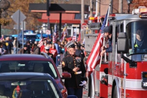 Man in uniform looking up at man holding American flag while leaning out of a fire truck window.