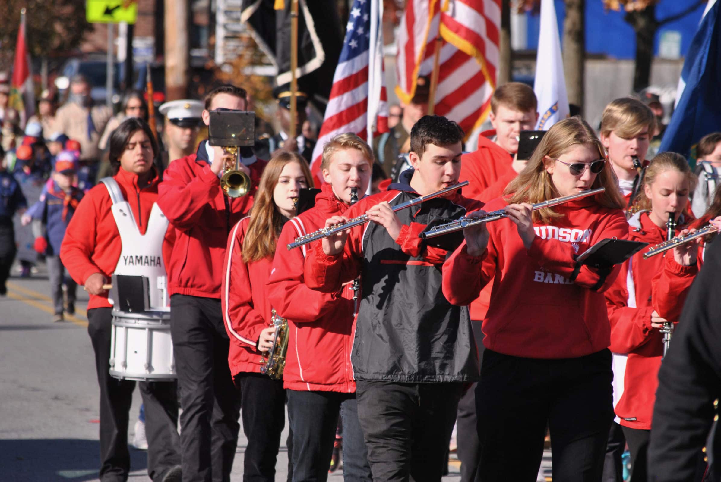 Students in a marching band wearing red jackets. Some carrying American flags.