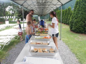 Members of Westboro Tennis & Swim Club line up at the barbecue to choose from freshly grilled hamburgers and hot dogs, fruits and vegetables, a pasta dish, watermelon, and chips.