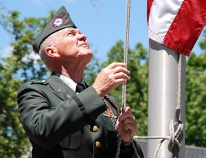 Parade Coordinator Bruce Goldsmith raises the American flag at the World War II – Korean War – Vietnam War Memorial.