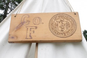 A wooden block engraved with the 13th Regiment Massachusetts Volunteers logo hangs above a tent.